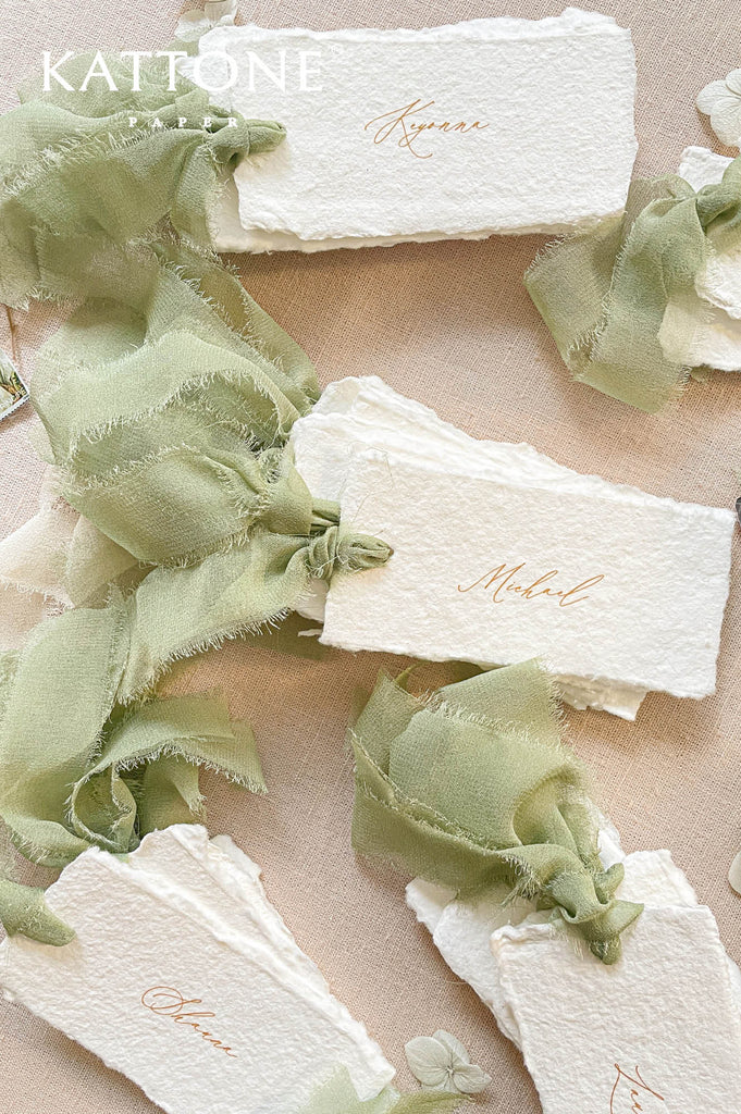 Overhead close-up of luxury wedding place cards made from ivory handmade deckled-edge paper, personalized with elegant warm script names and tied with soft sage green silk ribbon, arranged on natural linen with delicate botanical accents
