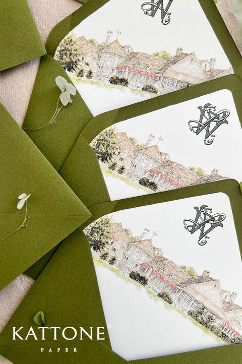 Three Kattone Paper stationery cards with a watercolor illustration of a village scene, arranged with olive green envelopes on a neutral fabric background.