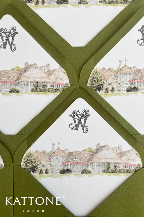 Angled overhead detail view of custom wedding envelope liners featuring a watercolor country club estate illustration, intertwined monogram, olive green envelopes, red architectural accents, and greenery