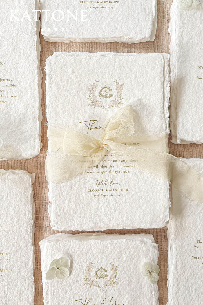 Close-up flat lay of a handmade paper wedding thank you card with deckled edges, soft gold typography, and a delicate floral crest, styled on neutral linen