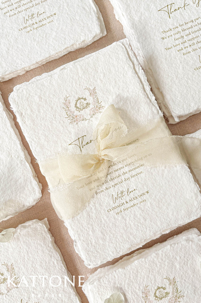 Top-down flat lay of multiple handmade paper thank you cards with deckled edges, floral crest details, and a soft ivory chiffon ribbon wrapped around the center card