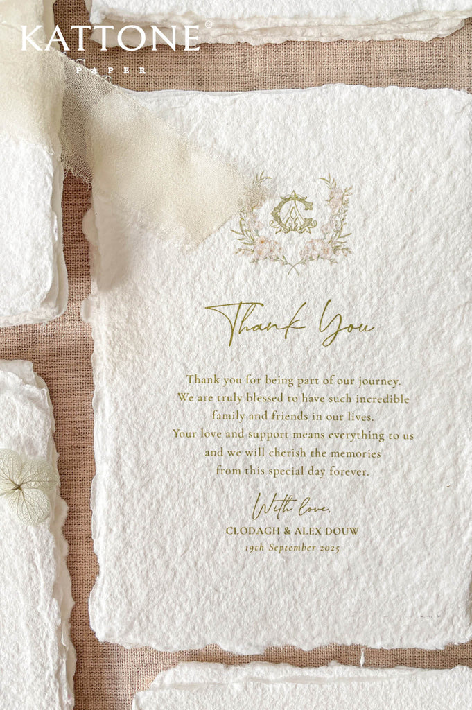 Angled overhead close-up of a textured handmade paper thank you card with deckled edges, romantic floral crest, elegant gold lettering, and a sheer ivory ribbon bow