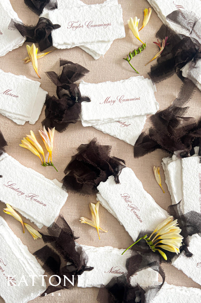 Top-down flat lay of personalized handmade paper wedding place cards with deckled edges, each printed with guest names in soft brown calligraphy, styled across neutral linen with sheer black ribbon and scattered yellow flowers for an elegant reception table setting display