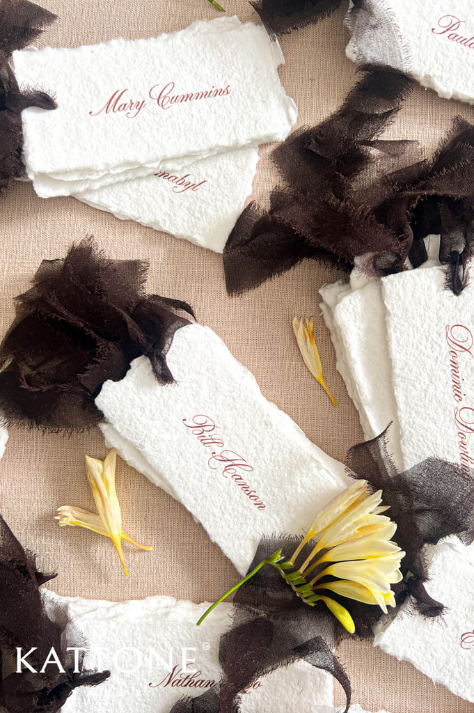White textured place cards with dark brown tulle accents and embroidered names, arranged on a beige fabric background with yellow flowers. 