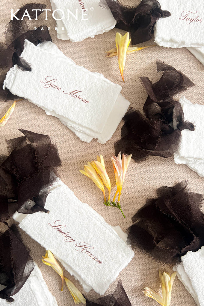White paper place cards with elegant cursive script names, styled with dark brown sheer ribbon and yellow flowers on a beige fabric background. 