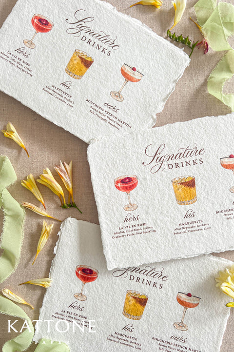 Top-down flat lay of handmade deckled-edge signature drinks wedding menus displayed on a neutral linen background, featuring watercolor cocktail illustrations, elegant burgundy script lettering, soft serif text, scattered yellow flowers, and pale green silk ribbon, styled as romantic custom wedding bar menu stationery