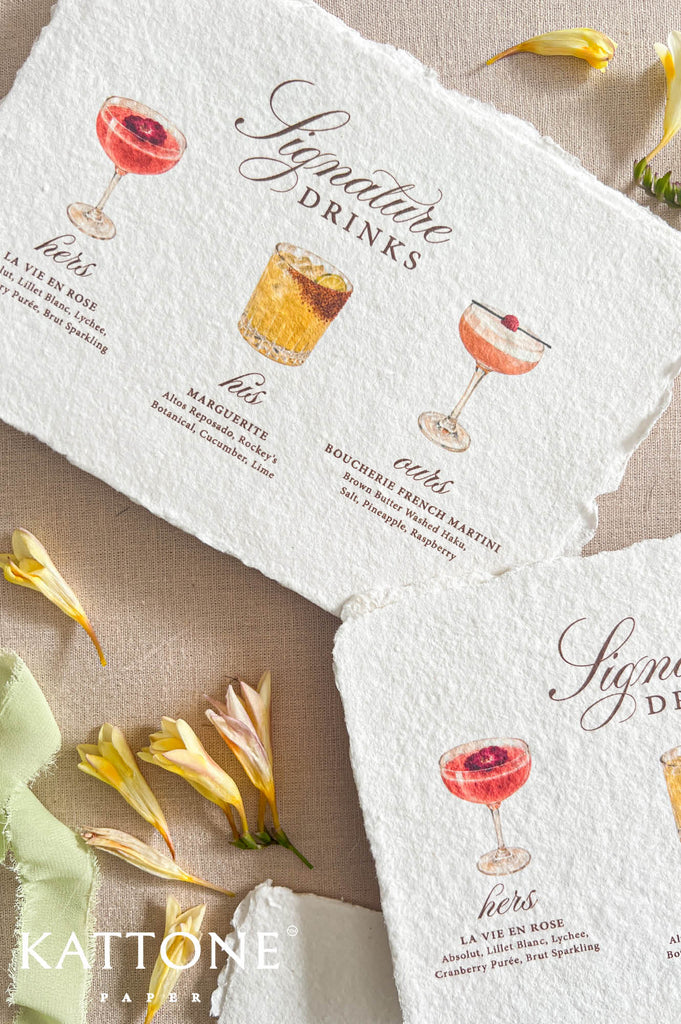 Two rustic white paper napkins with printed cocktail illustrations and the words 'Signature Drinks' are arranged on a beige fabric surface with yellow flower petals. The brand name Kattone Paper appears in the lower left corner.