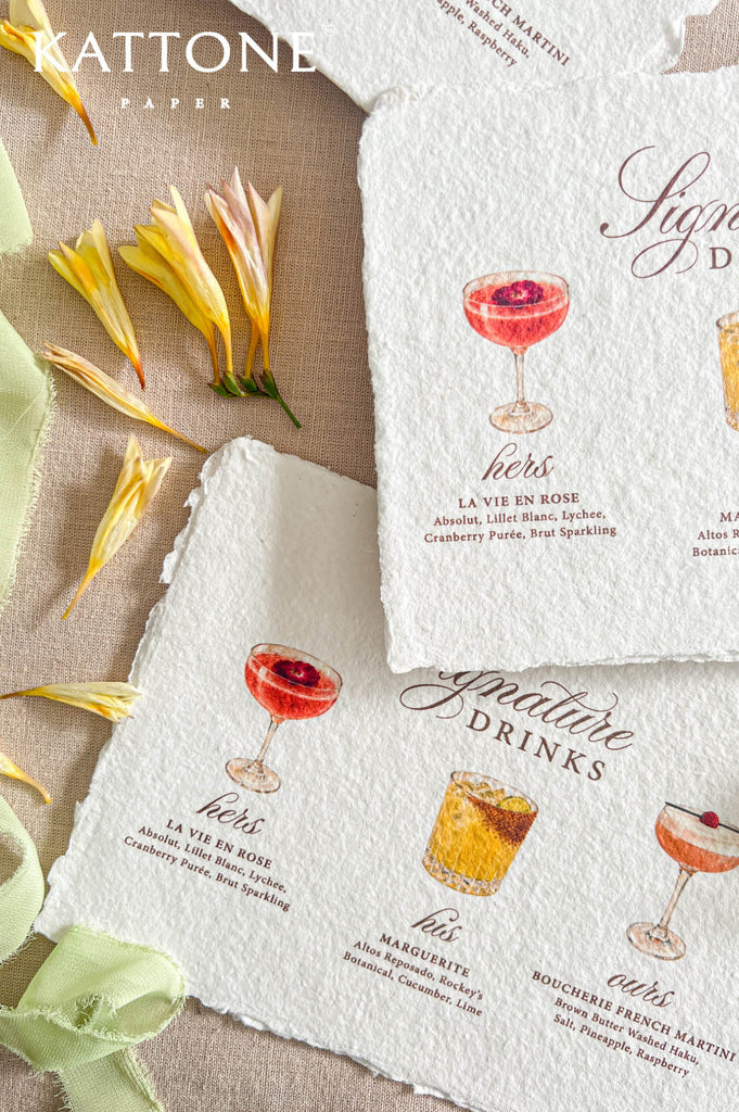 Two textured cream paper napkins with printed cocktail illustrations and French text, arranged on a beige fabric background with yellow flower petals and a green ribbon. The brand name KATTONE PAPER appears in the top left.