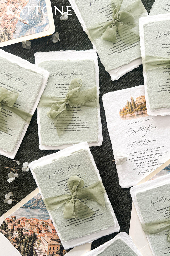 A styled flat-lay of wedding invitation cards with textured off-white paper, pale sage green ribbons tied in bows, and printed event details, arranged on a dark fabric background with small floral props.