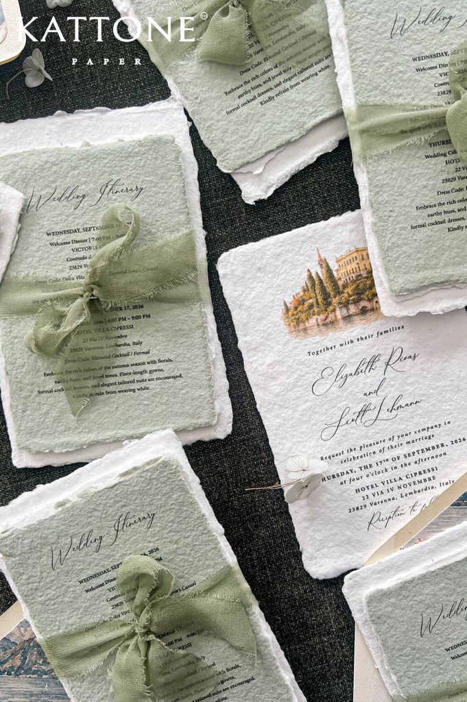 A styled flat-lay of Kattone Paper wedding stationery, featuring textured off-white and sage green cards tied with sage green silk ribbons, arranged on a dark charcoal fabric background.