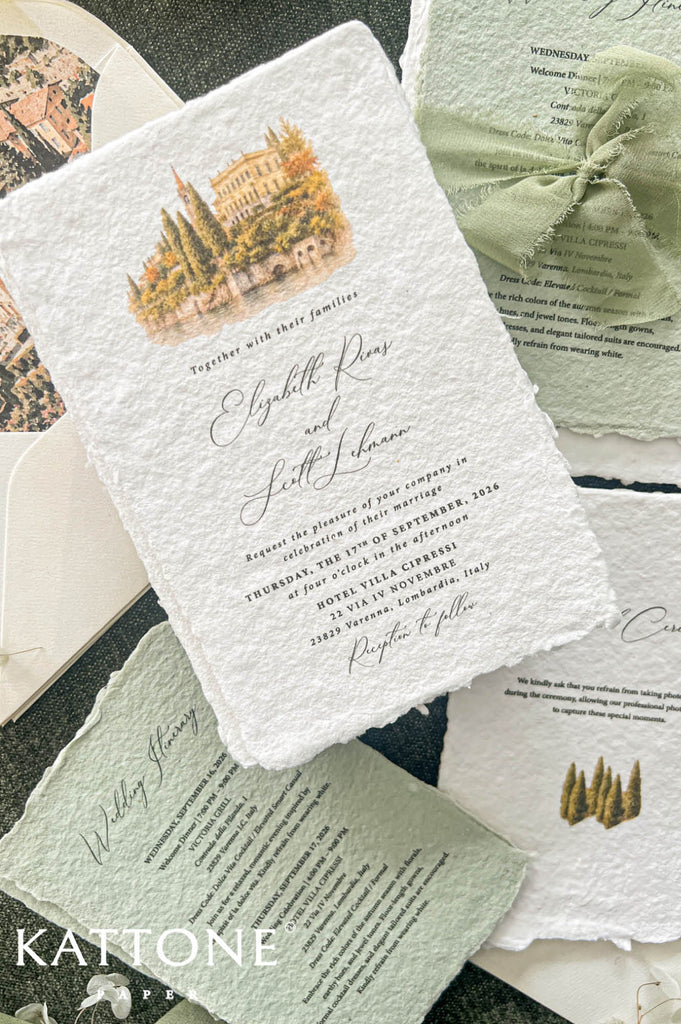 A styled flat-lay of wedding stationery from Kattone, featuring textured handmade paper cards with a rustic watercolor illustration of a villa and cypress trees, a sage green ribbon bow, and matching envelopes on a dark textured background.