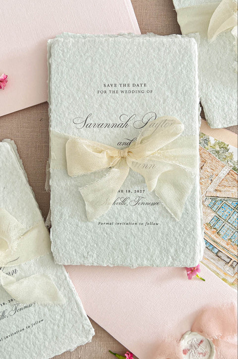 Close-up flat lay of colored handmade paper save the date card with deckled edges and an ivory ribbon bow, styled beside a blush envelope with a custom architectural illustration liner