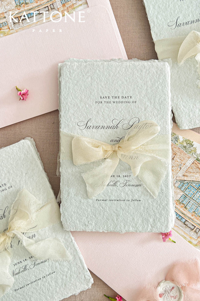 Close-up flat lay of colored handmade paper save the date card with deckled edges and an ivory ribbon bow, styled beside a blush envelope with a custom architectural illustration liner