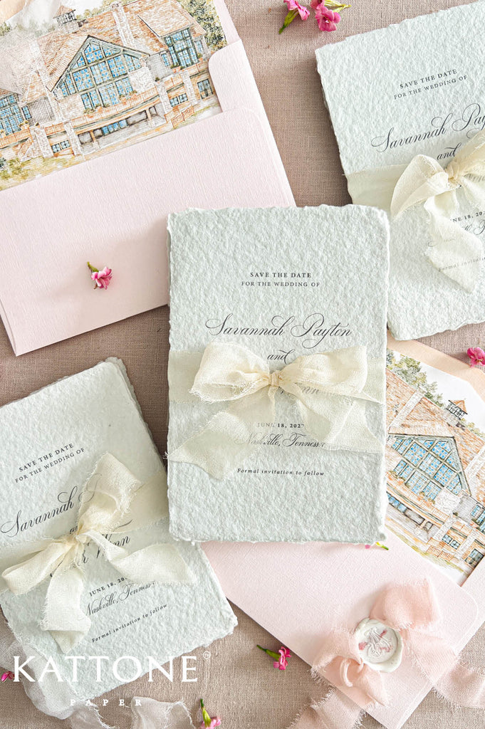 A styled flat lay of wedding stationery from Kattone Paper, featuring textured pale green and blush pink cards tied with cream satin ribbons, a matching envelope with a house illustration, and small pink flowers on a neutral fabric background.