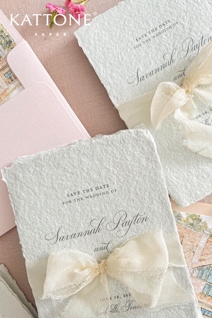 Two textured off-white wedding save-the-date cards tied with cream lace bows, arranged on a blush pink background with a pink envelope and a small floral accent. The brand name Kattone Paper appears in the top left.