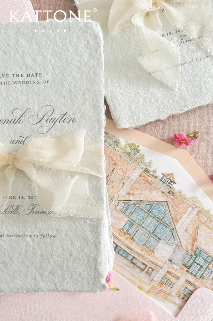 Top-down flat lay of multiple colored handmade paper save the date cards with soft ivory ribbon bows, blush pink envelopes, custom illustrated envelope liners, and delicate floral accents on neutral linen