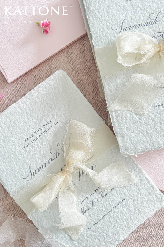 Two textured white wedding invitation cards tied with cream ribbon bows, arranged on a soft pink surface with a pink envelope and small flower accent. The brand name KATTONE FAUX is visible in the top left.