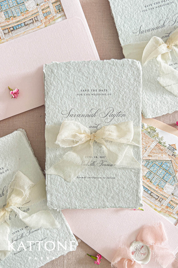 A styled flat-lay of wedding stationery featuring a central textured pale blue-green save-the-date card tied with a cream ribbon bow, surrounded by matching cards, envelopes, and pink paper accents on a beige surface.