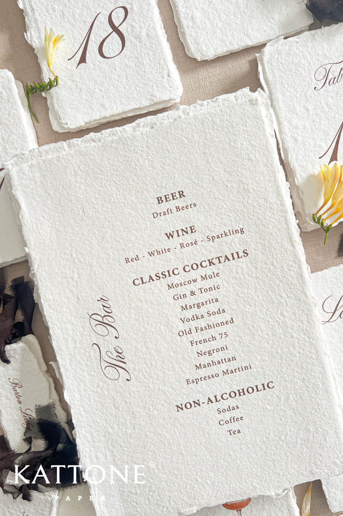 A styled flat-lay of rustic white paper place cards with rough deckled edges, printed with event details like a beverage list and table number 18, accented with small yellow flowers on a neutral beige surface. The brand name KATTONE EVENTS appears in the lower left.
