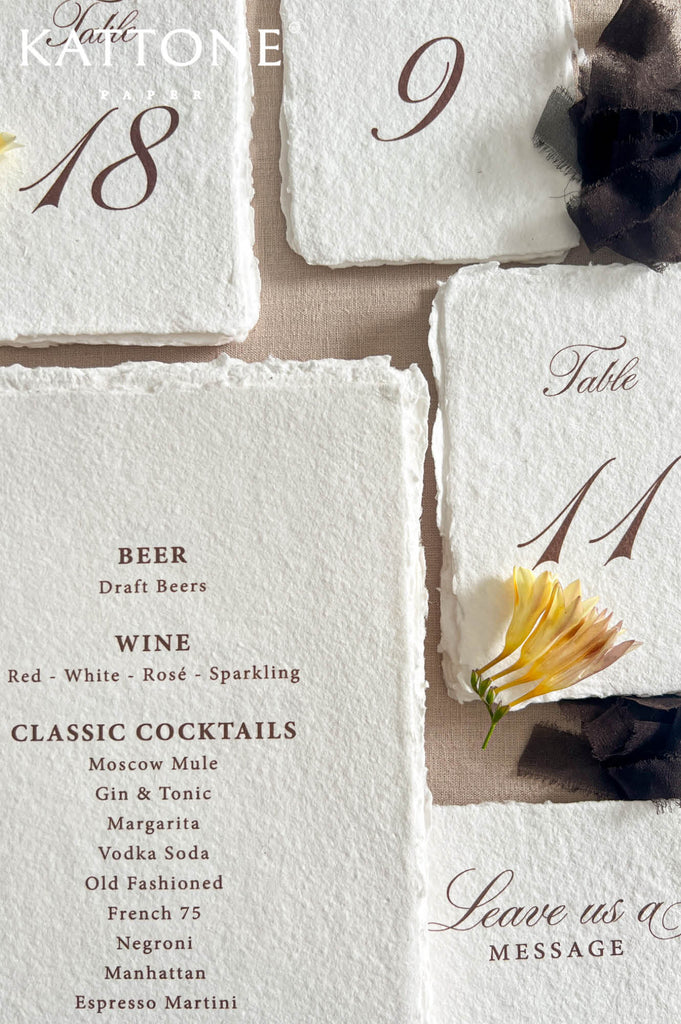 Overhead close-up of elegant handmade paper wedding bar menu with deckled edges, burgundy script title, organized beer, wine, cocktail, and non-alcoholic drink list, styled among ivory table numbers, cocktail illustrations, dark ribbon, and yellow flowers