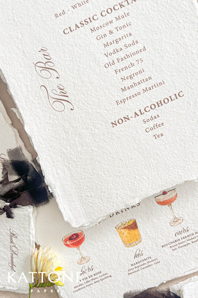 A styled flat-lay of a textured off-white menu card with elegant copper-brown typography, partially layered over another printed card featuring illustrated cocktails. The composition includes decorative paper edges, a small yellow flower, and a dark feather on a light neutral surface.