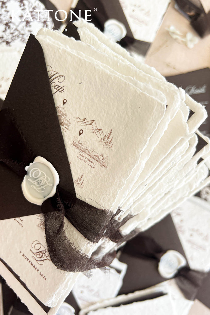A close-up of a stack of textured cream-colored paper cards or envelopes tied with a sheer black ribbon and a white wax seal, with printed script and map-like illustrations. The brand name ATONE is visible in the top left corner.