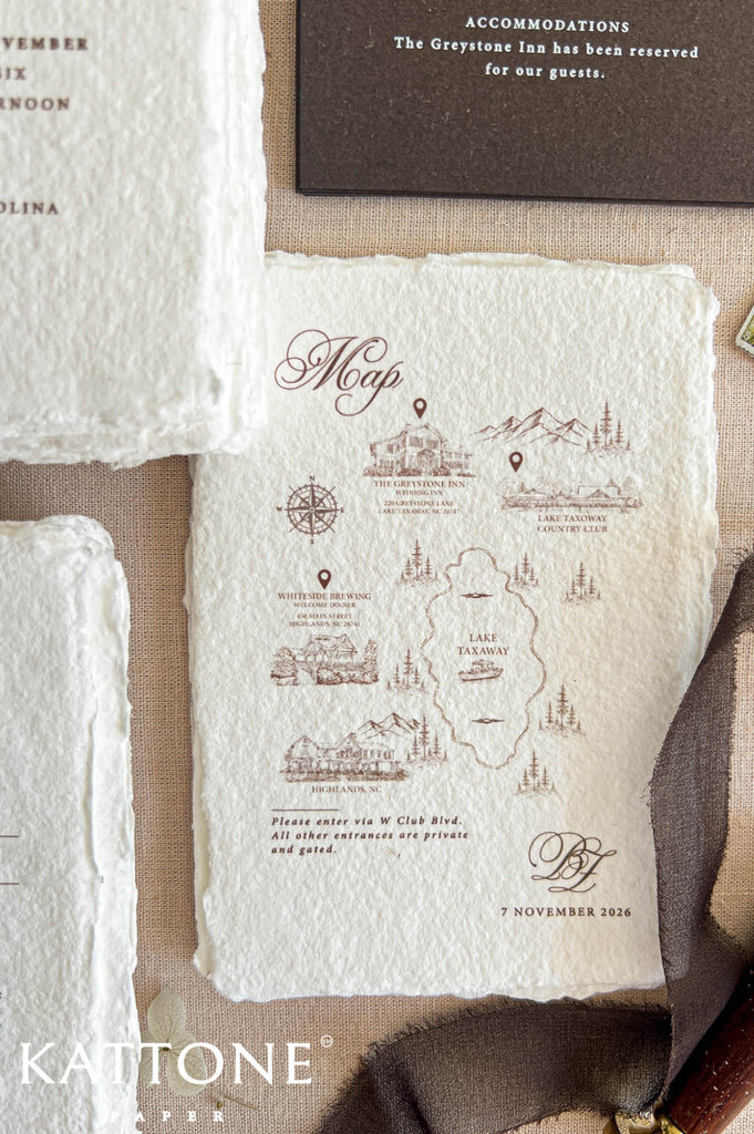 A styled flat-lay of vintage-style paper stationery, with a central cream-colored textured paper card featuring brown illustrated map details and elegant script. Partially visible coordinating papers and a dark brown envelope surround it on a neutral fabric background.