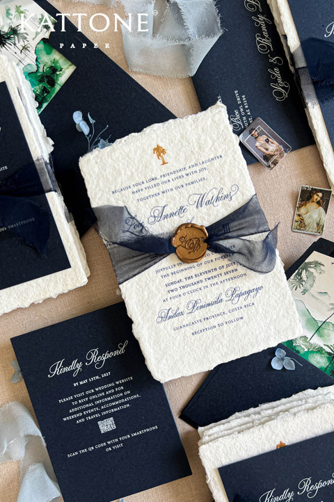 A styled flat-lay of wedding stationery from Kattone Papers, featuring a textured cream invitation envelope tied with a navy ribbon and gold wax seal, surrounded by navy and cream cards, photos, and decorative elements on a neutral background.