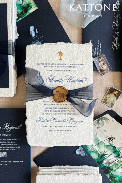A styled flat-lay of wedding stationery from Kattone, featuring a cream textured invitation envelope tied with a navy ribbon and a gold wax seal, alongside matching navy cards, a floral green-and-white napkin, and small printed photos on a neutral surface.