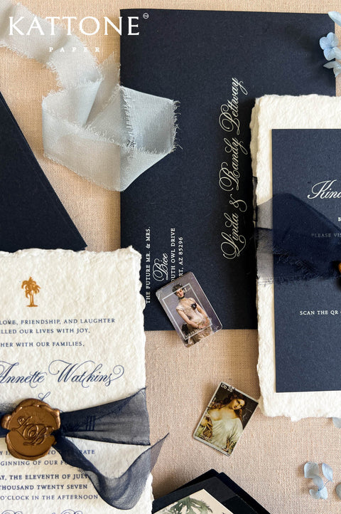 A styled flat-lay of a Kattone by Kattone wedding invitation suite on a beige fabric background. The suite includes a dark navy blue cardstock invitation with gold foil script, a cream textured paper invitation with blue script, a navy ribbon, a gold wax seal, and small photo inserts.