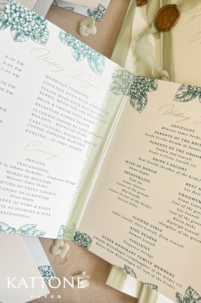 An open wedding program booklet with printed event program text, decorated with teal botanical leaf borders and gold script headings, styled on a beige fabric surface with a wax seal and small white beads.