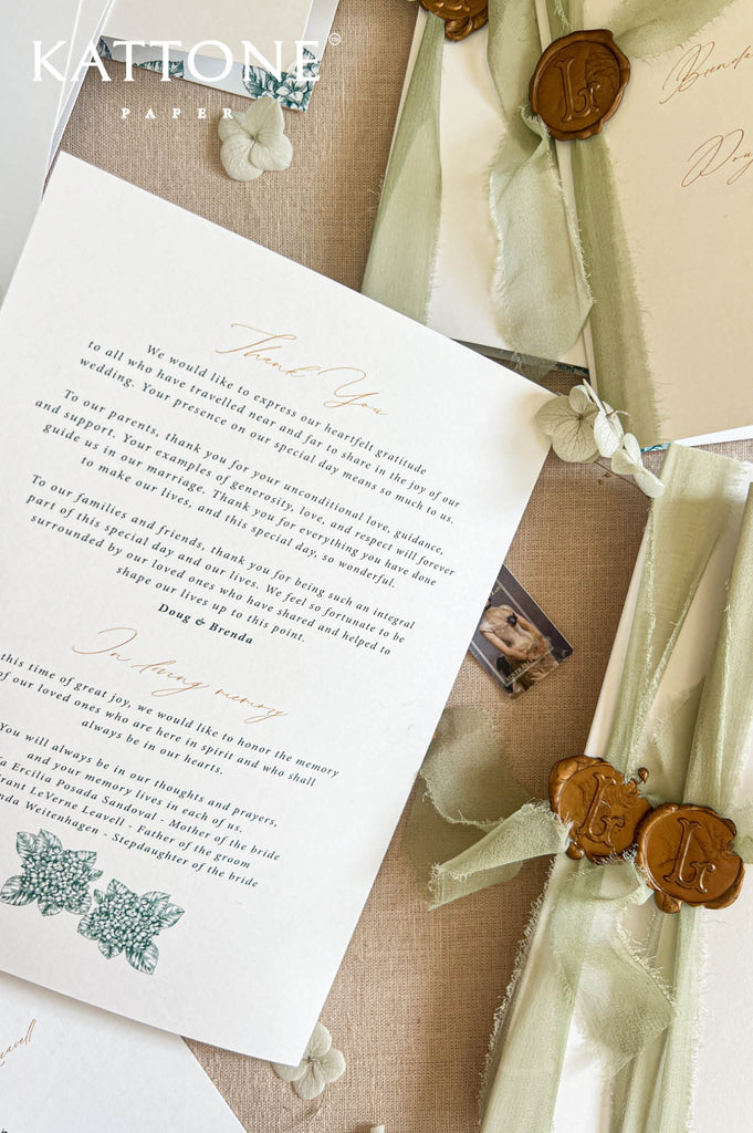 Overhead close-up of opened folded wedding programs showing ceremony timeline and wedding party details, with green botanical border illustrations, refined typography, and coordinating stationery layered on a linen backdrop