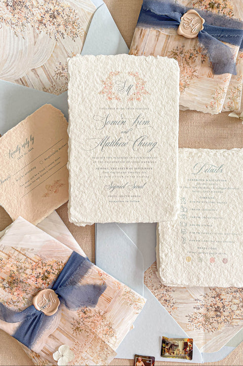 A styled flat-lay of a wedding invitation suite from Kattone, featuring textured cream paper, floral watercolor designs, dusty blue ribbons, and gold wax seals on a neutral beige background.
