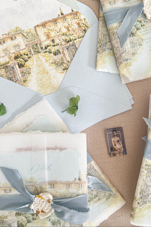 A styled flat-lay of coordinated stationery sets featuring watercolor villa illustrations, pale blue envelopes, translucent paper, ribbon bows, and decorative wax seals on a beige fabric background.
