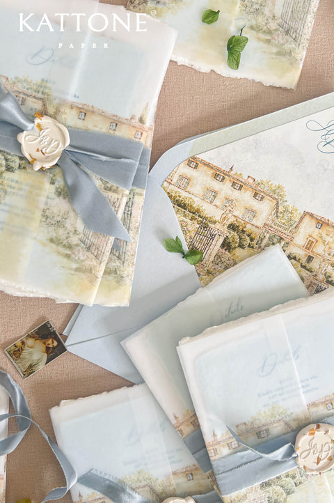 A styled flat-lay of elegant stationery from Kattone Paper, featuring printed villa illustrations, light blue ribbons, wax seals, and matching envelopes on a beige fabric background.