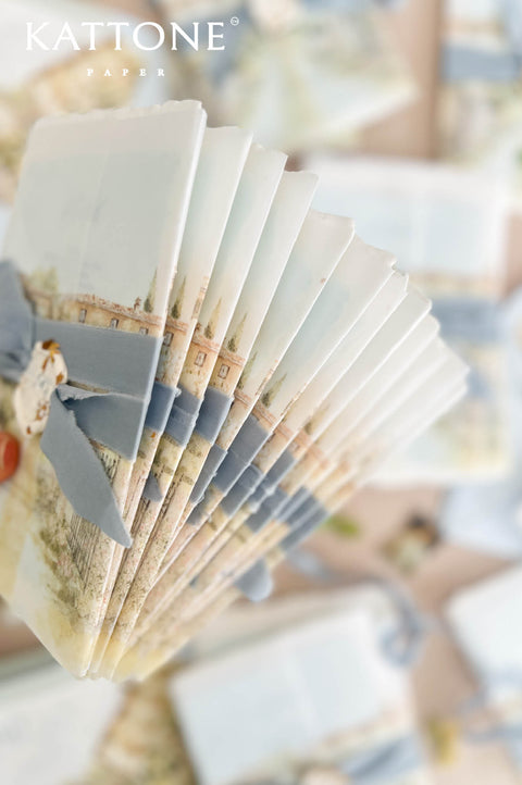 A fanned stack of Kattone Paper postcards decorated with a dusty blue ribbon and a small white tag, set against a soft-focus background of more postcards.