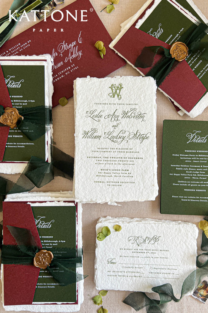 Top-down flat lay of luxury burgundy and forest green wedding invitation suite featuring ivory handmade deckled-edge paper, elegant calligraphy, burgundy envelopes, green details cards, dark sheer ribbon, antique-gold monogram wax seals, and coordinating RSVP stationery