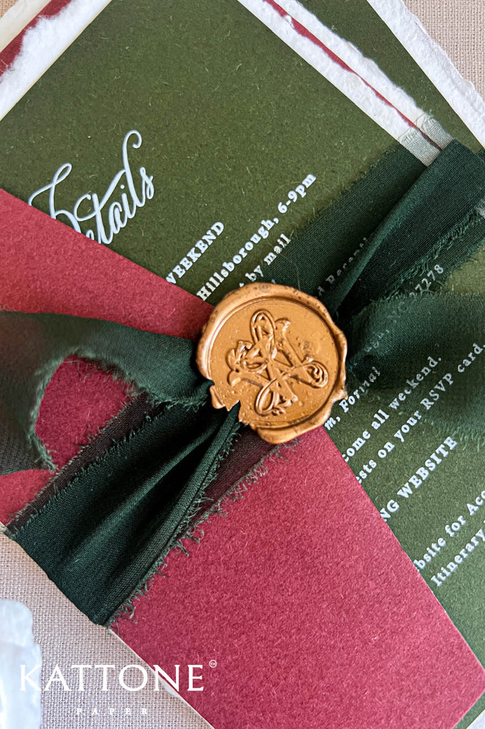 A close-up of a Kattone invitation card featuring a dark green background with white script and block text, layered with a deep red paper ribbon and tied with a dark green satin ribbon. A gold wax seal with an embossed monogram is centered on the bow.