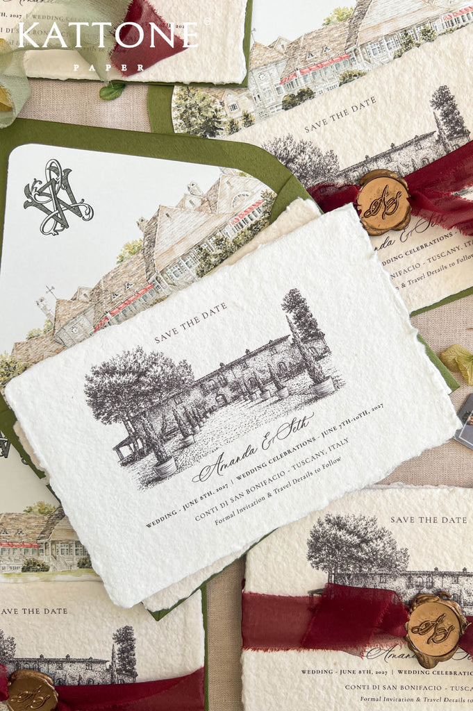Angled overhead close-up of luxury handmade paper save the date card wrapped with burgundy ribbon and personalized bronze wax seal, layered over an olive green envelope with custom watercolor venue liner and monogram artwork