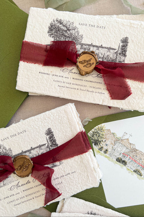 Top-down flat lay of handmade deckled-edge save the date cards featuring black architectural venue illustrations, deep burgundy sheer ribbon, bronze monogram wax seals, olive green envelopes, and watercolor envelope liners styled on neutral linen