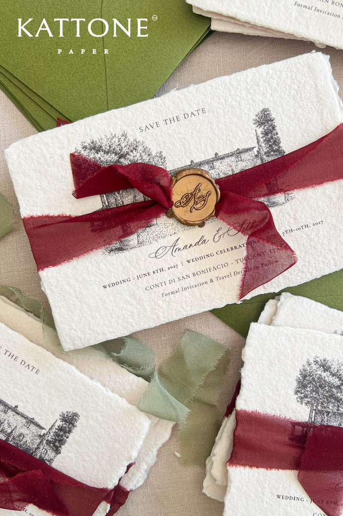 Kattone Paper 'Save the Date' cards featuring a textured off-white paper background with a black ink architectural illustration, tied with a deep burgundy ribbon and a round wooden wax seal. The cards are arranged with other matching stationery pieces on a neutral beige surface.