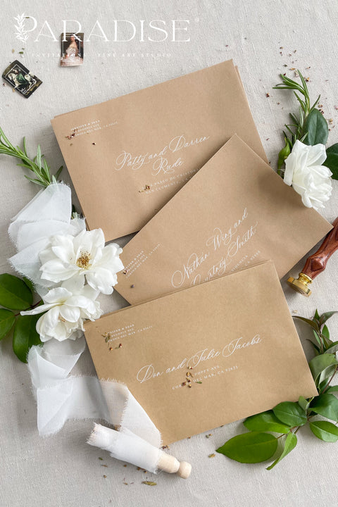 Neutral kraft wedding envelopes with white ink address printing, styled flat lay with white florals, greenery, and wax seal for minimalist and botanical invitation suites.
