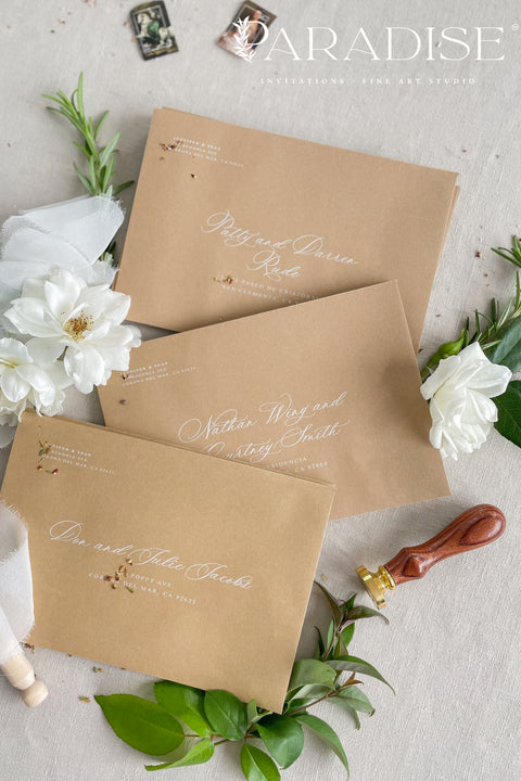 Neutral kraft wedding envelopes with white ink address printing, styled flat lay with white florals, greenery, and wax seal for minimalist and botanical invitation suites.