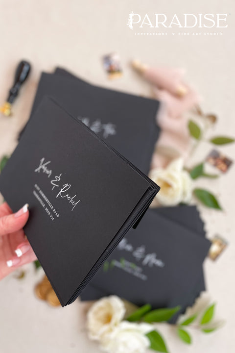 Matte black wedding envelopes with white ink address printing, styled flat lay with florals, wax seal, and greenery, perfect for modern and editorial invitation suites.