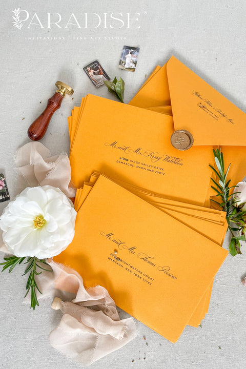 Amber wedding envelopes with printed addresses, styled flat lay with white florals, wax seal, ribbon, and greenery, perfect for warm, elegant invitation suites.