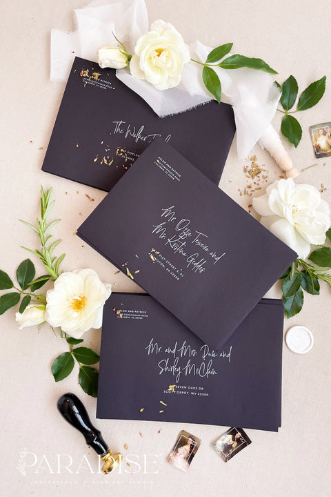 Modern black envelopes with white ink calligraphy printing, photographed flat lay with white flowers, wax seal, and botanical details.