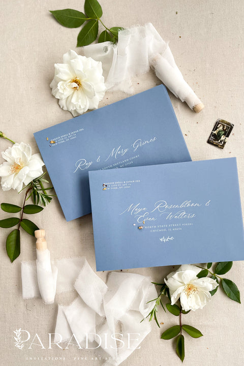 Dusty blue wedding envelopes with printed addresses, styled flat lay with white florals, ribbon, and greenery, perfect for elegant and coastal invitation suites.