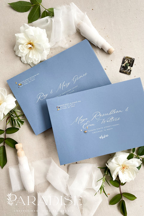 Dusty blue wedding envelopes with printed addresses, styled flat lay with white florals, ribbon, and greenery, perfect for elegant and coastal invitation suites.