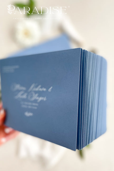 Dusty blue wedding envelopes with printed addresses, styled flat lay with white florals, ribbon, and greenery, perfect for elegant and coastal invitation suites.