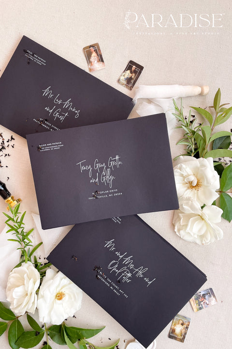 Modern black envelopes with white ink calligraphy printing, photographed flat lay with white flowers, wax seal, and botanical details.